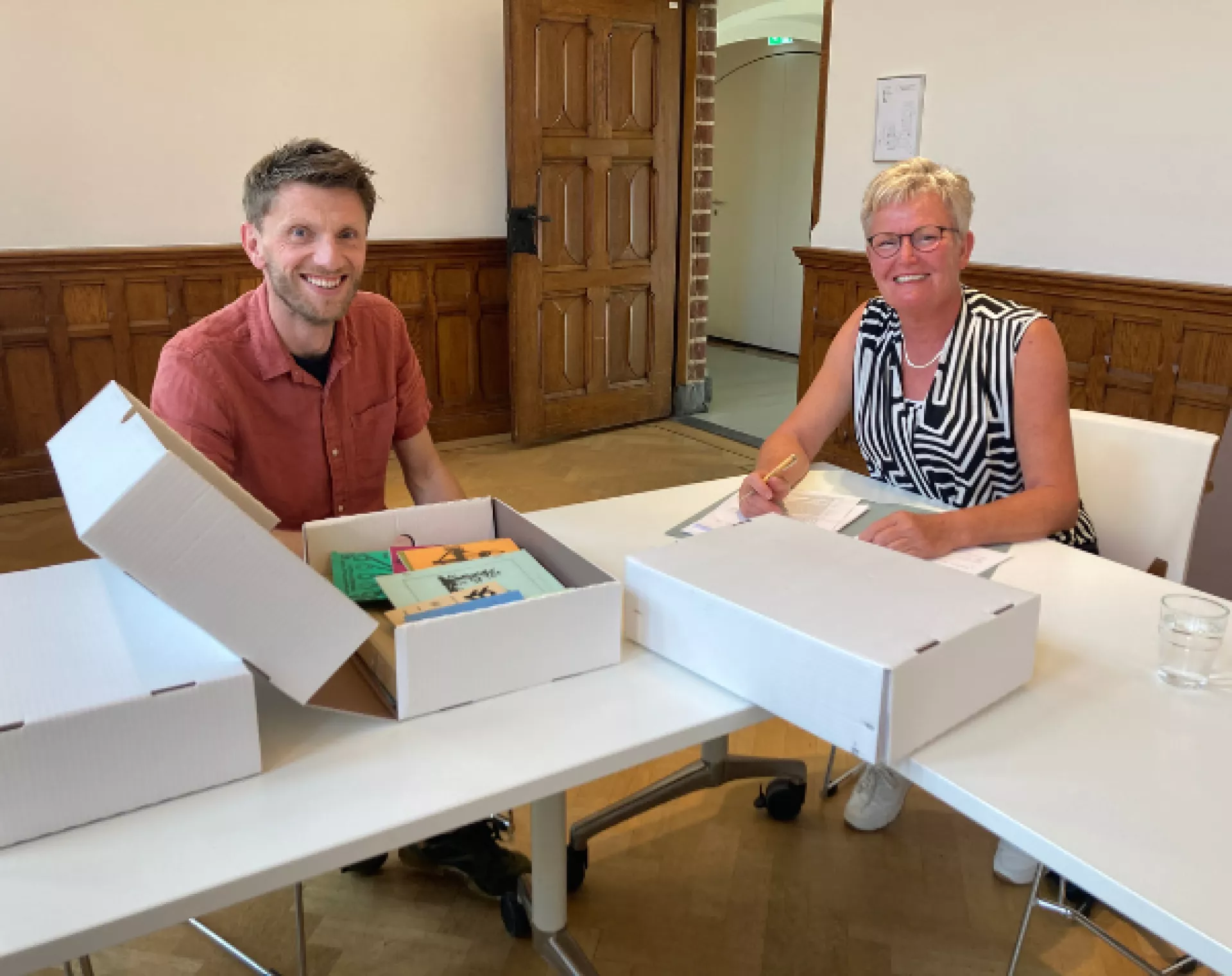 Man en vrouw in zaal, kijken lachend in de camera. Op tafel liggen archiefdozen met archiefmateriaal.