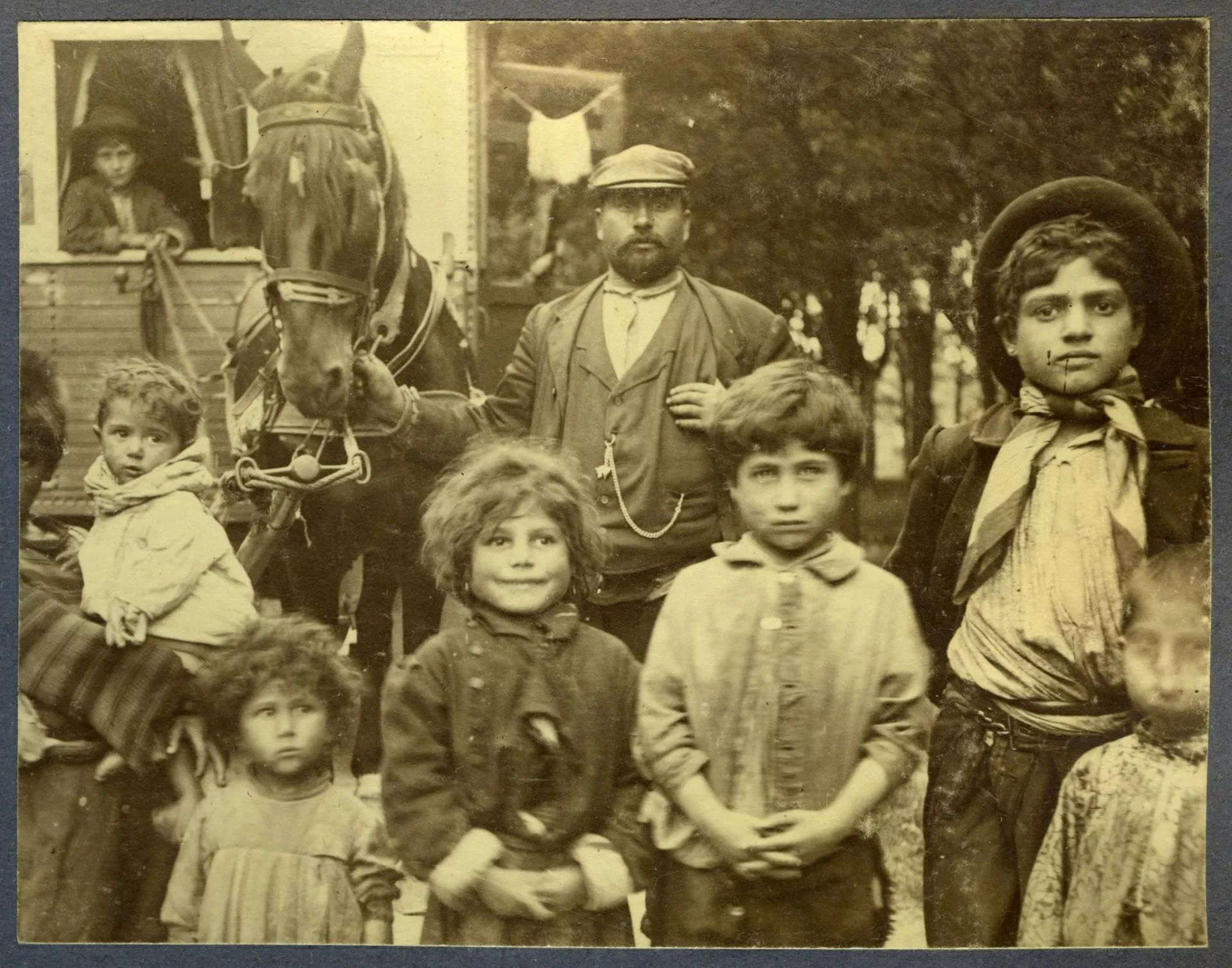 Zwart-wit foto groep reizigers. Man met pet staat in het midden. Eromheen kinderen in verschillende leeftijden. Man houdt paard vast met een kar daarachter.