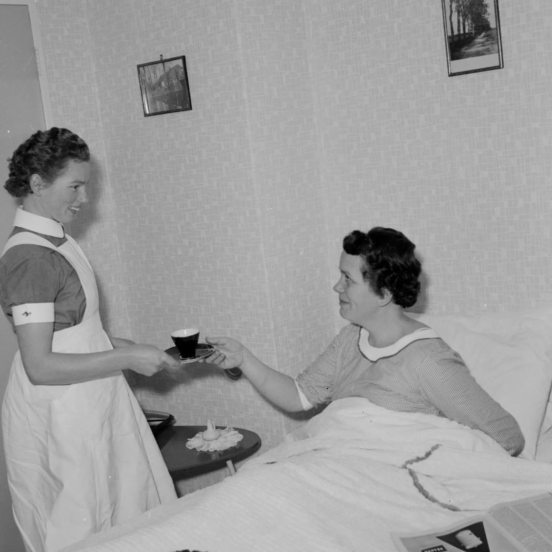 Zwart-wit foto. Verpleegster brengt vrouw in bed een kop koffie. Ze kijken elkaar glimlachend aan.