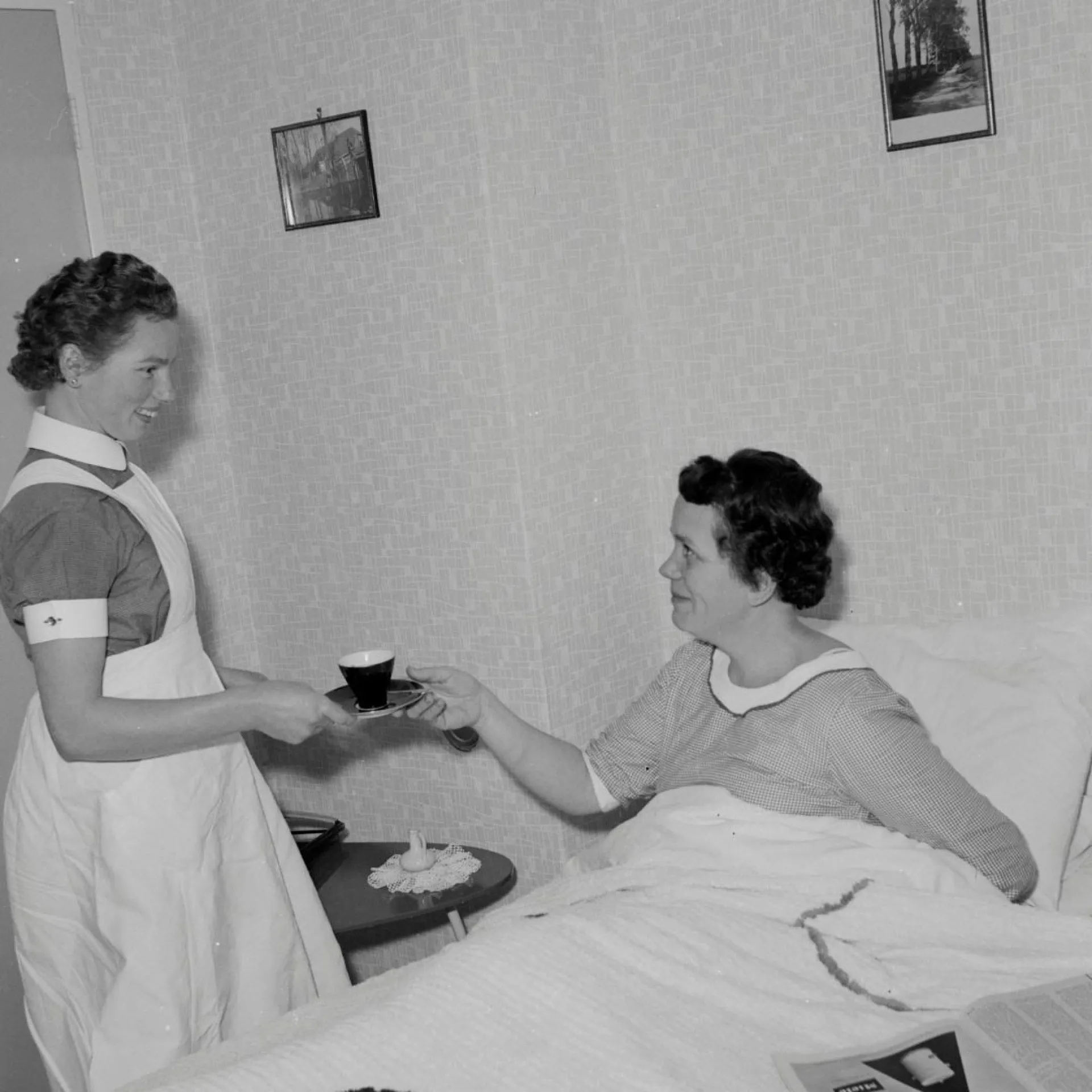 Zwart-wit foto. Verpleegster brengt vrouw in bed een kop koffie. Ze kijken elkaar glimlachend aan.