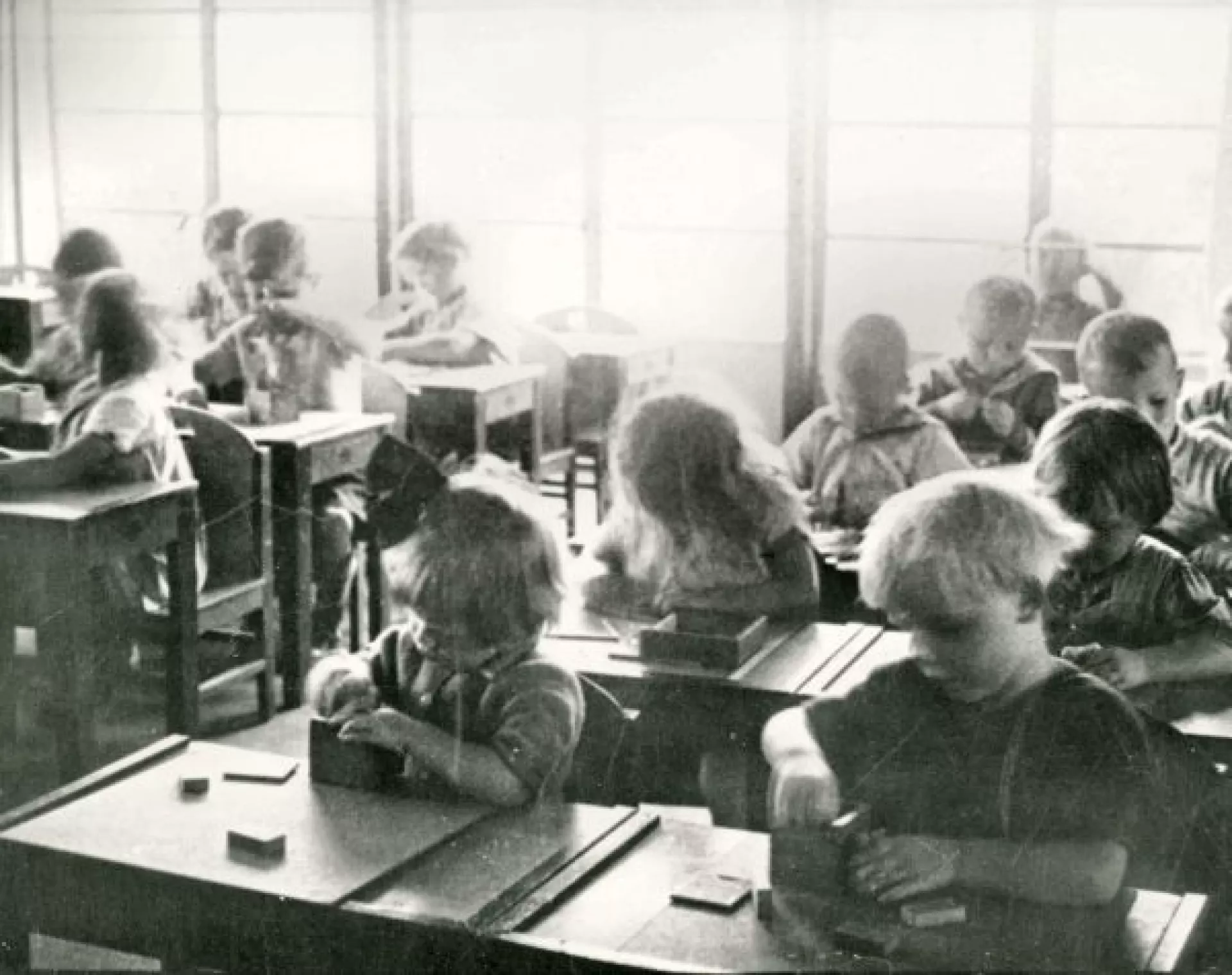 Zwartwit foto kleuterklas. We zien 15 kinderen van ongeveer 4 jaar oud. Vooraan zitten een jongetje en meisje die in opperste concentratie iets uit een houten doosje halen.