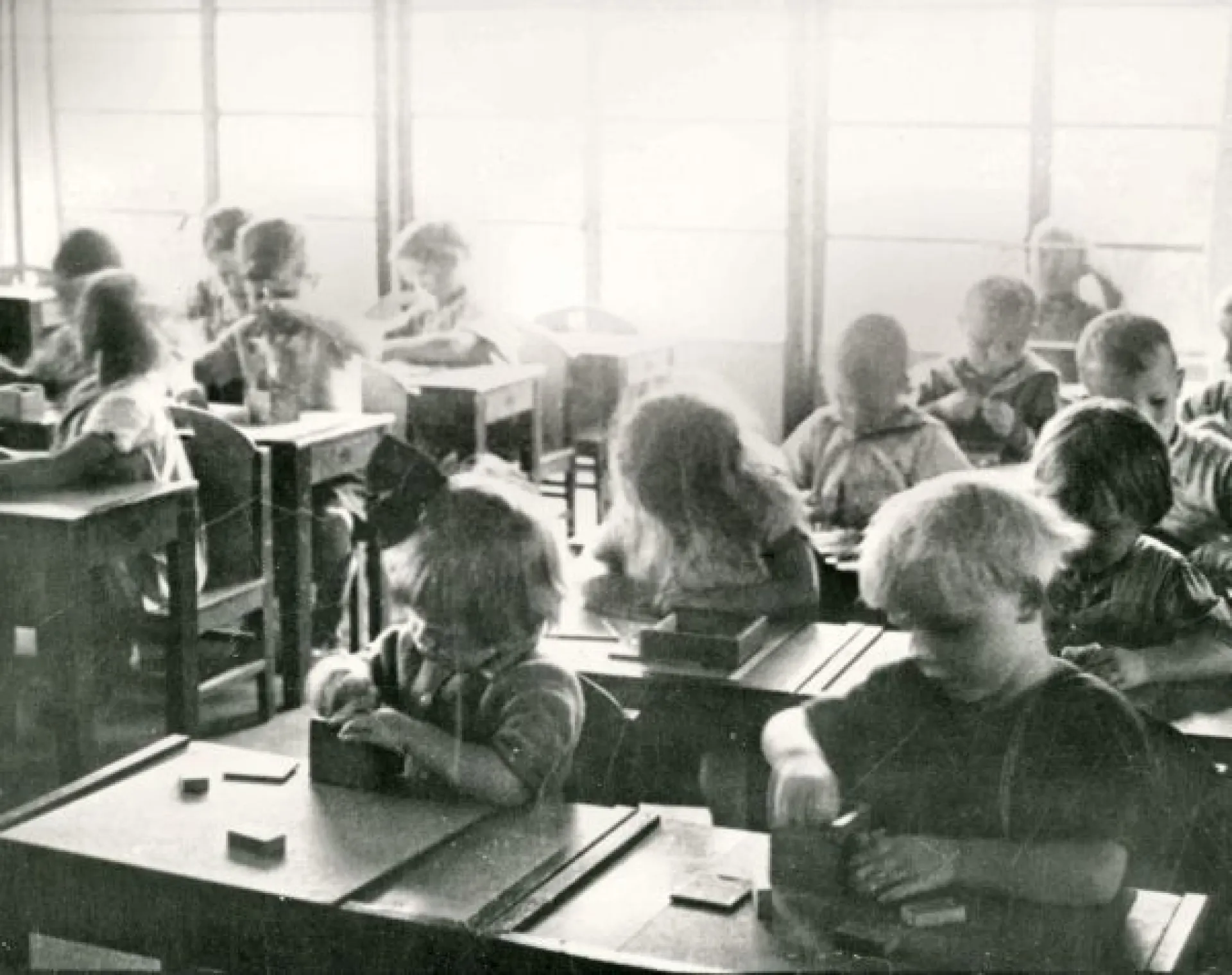 Zwartwit foto kleuterklas. We zien 15 kinderen van ongeveer 4 jaar oud. Vooraan zitten een jongetje en meisje die in opperste concentratie iets uit een houten doosje halen.