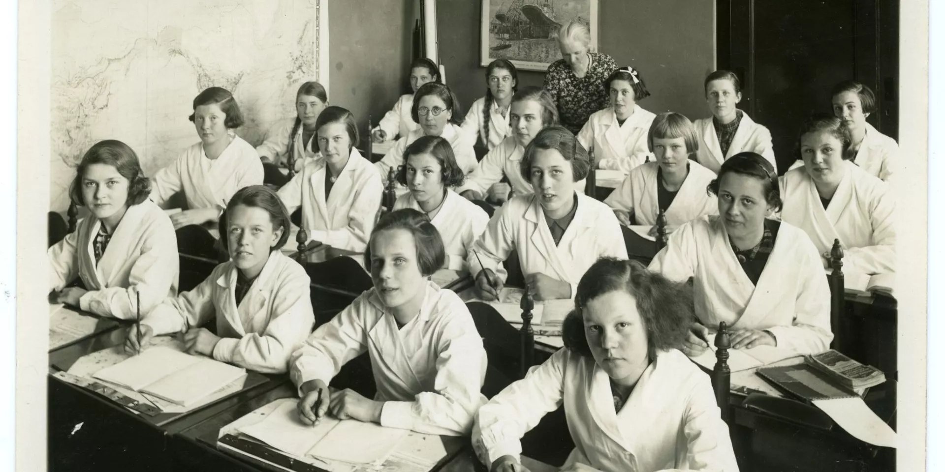 Klaslokaal met 20 meisjes en 1 juf. De leerlingen zitten aan tafel en schrijven. Ze dragen witte vesten. Op de muur hangt een landkaart en een afbeelding van een scheepswerf..