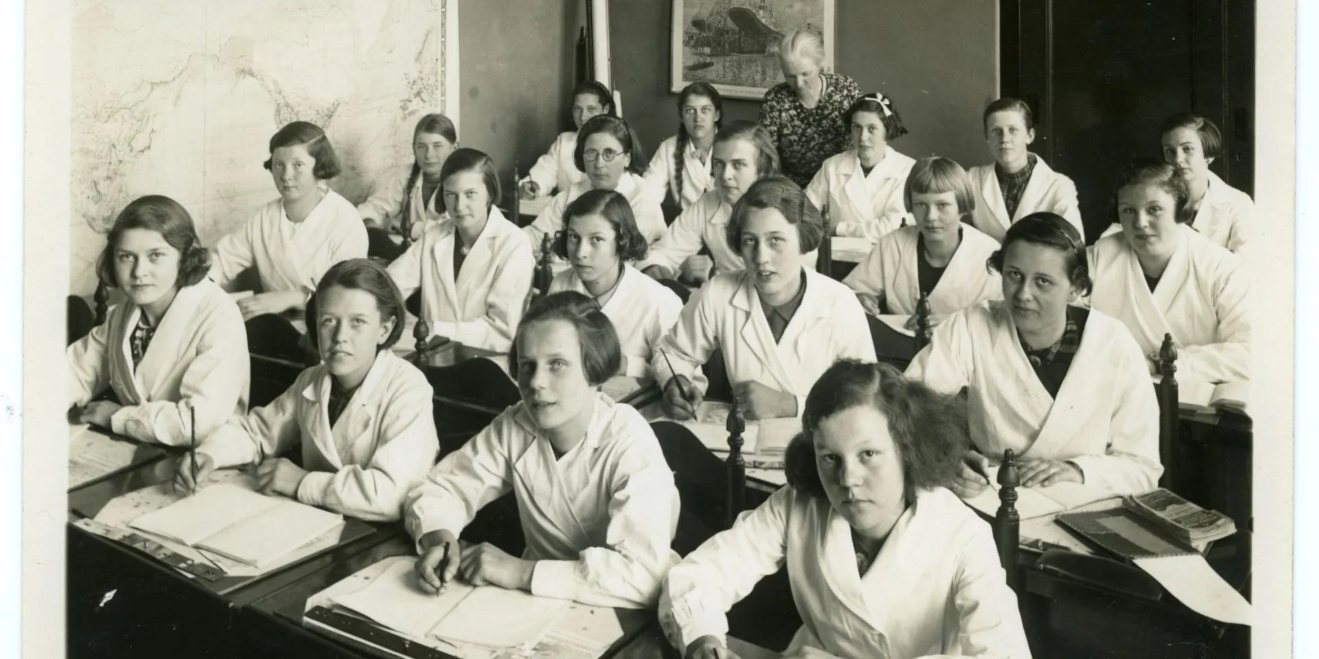 Klaslokaal met 20 meisjes en 1 juf. De leerlingen  zitten aan tafel en schrijven. Ze dragen witte vesten. Op de muur hangt een landkaart en een afbeelding van een scheepswerf..