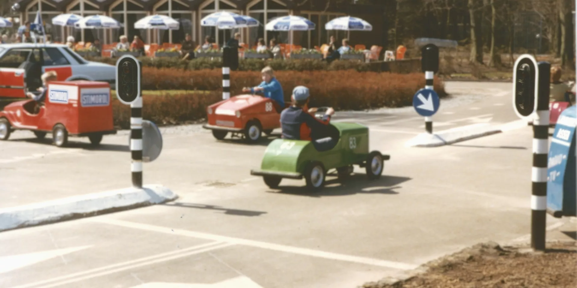Kleurenfoto van verkeerspark Assen. Er rijden drie kleine auto's, er staan verkeerslichten. Op de achtergrond zien we een terras met parasols.