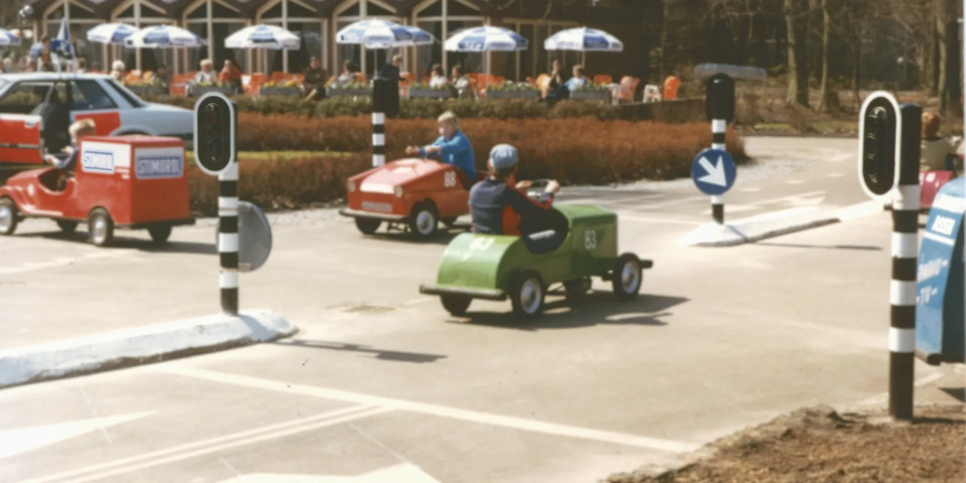 Kleurenfoto van verkeerspark Assen. Er rijden drie kleine auto's, er staan verkeerslichten. Op de achtergrond zien we een terras met parasols.