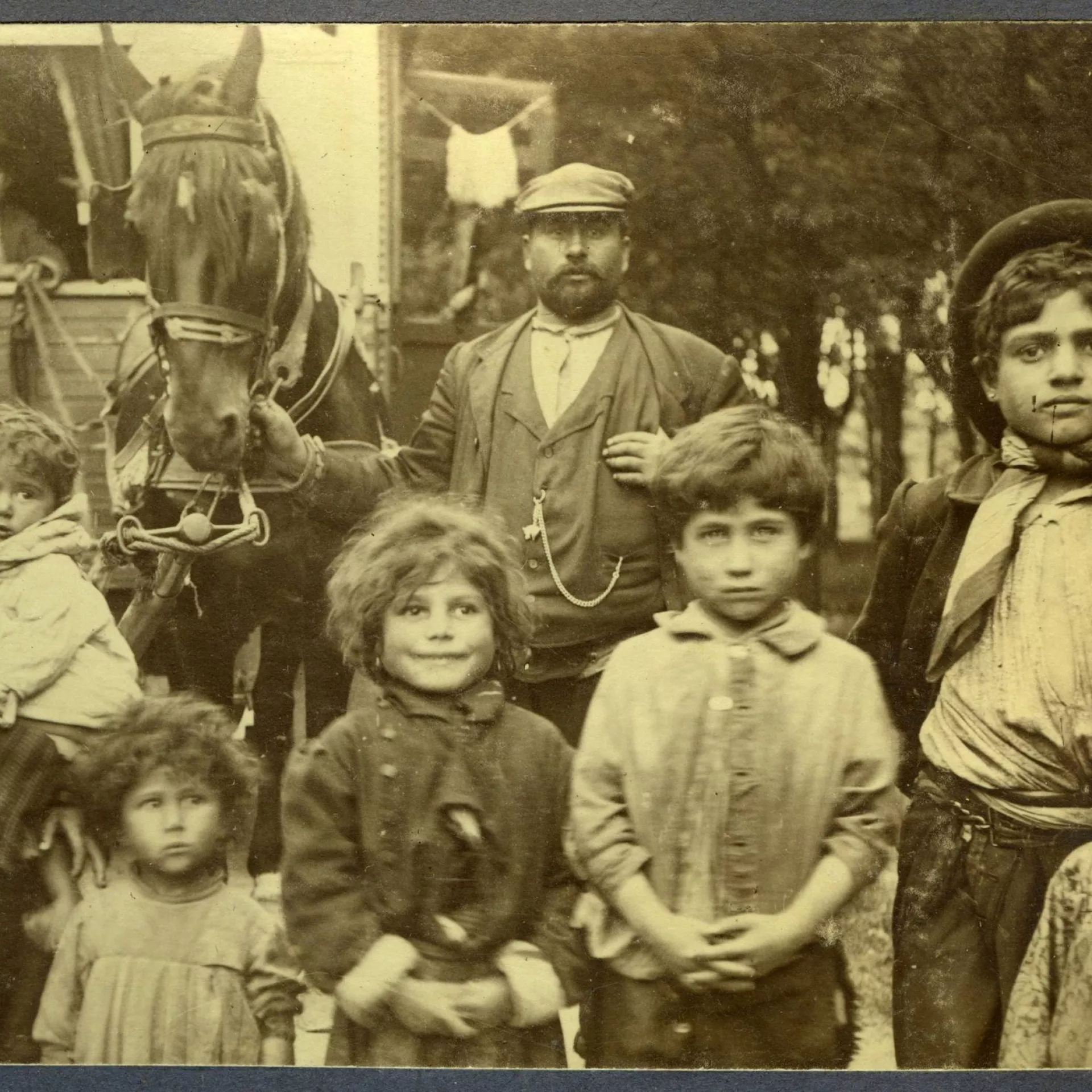 Zwart-wit foto groep reizigers. Man met pet staat in het midden. Eromheen kinderen in verschillende leeftijden. Man houdt paard vast met een kar daarachter.