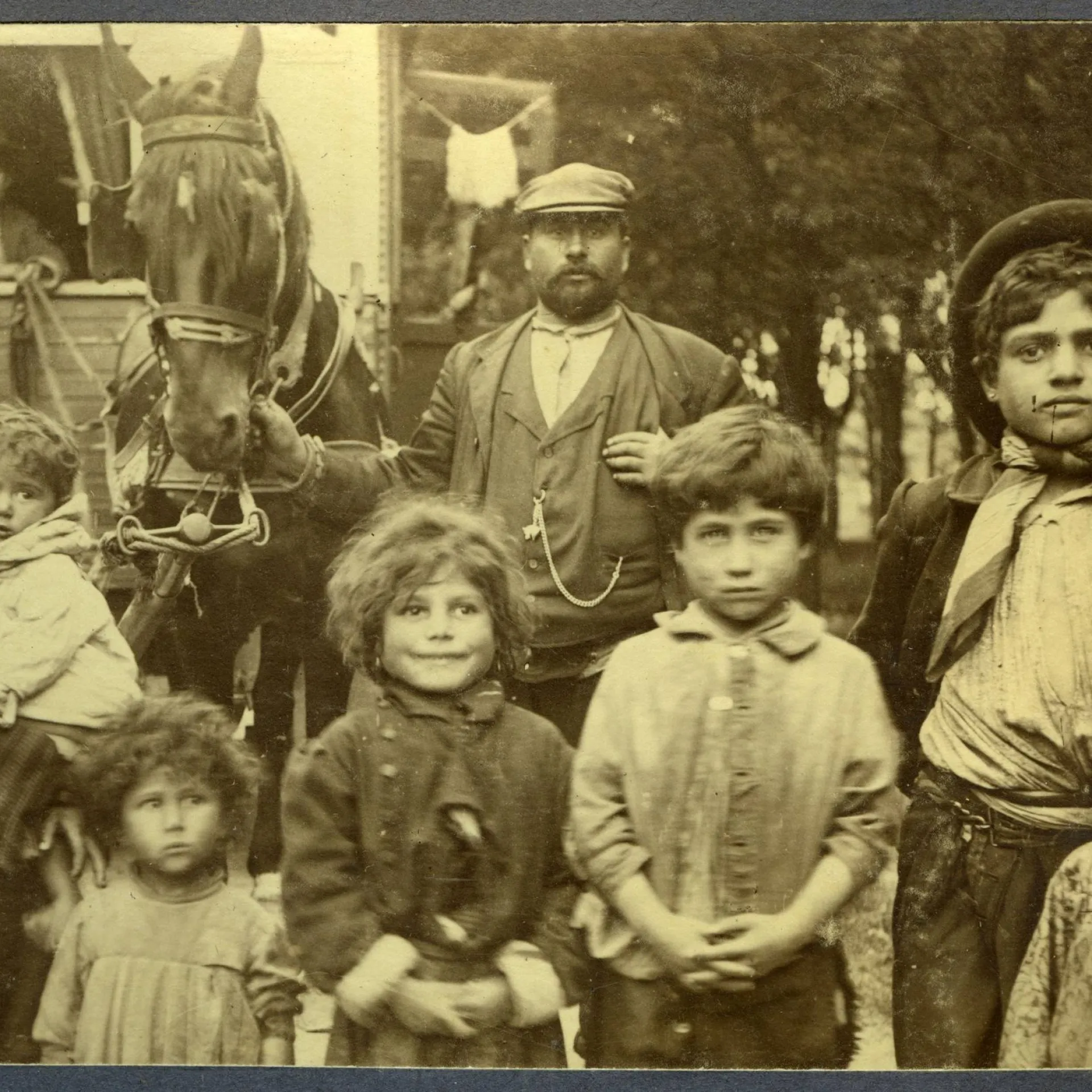 Zwart-wit foto groep reizigers. Man met pet staat in het midden. Eromheen kinderen in verschillende leeftijden. Man houdt paard vast met een kar daarachter.