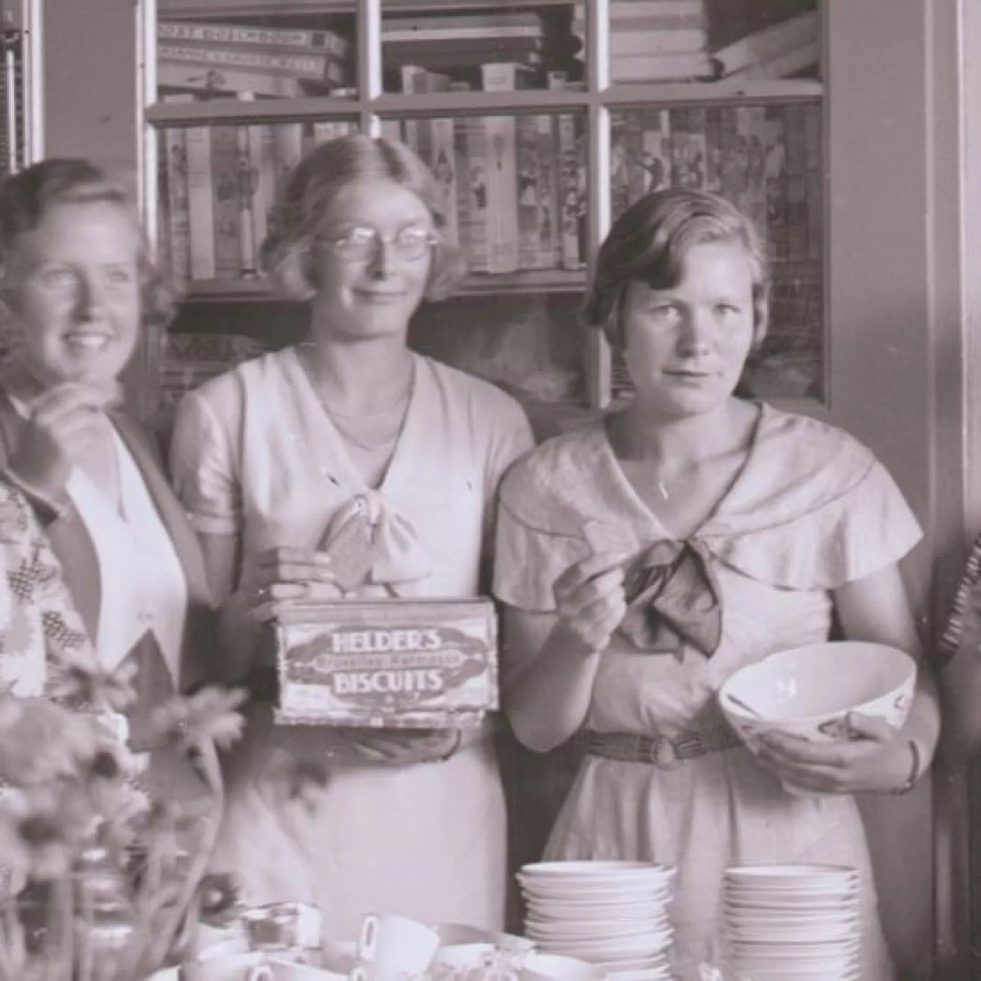 Zwart-wit foto. Groep van 8 vrouwen in klaslokaal, drinkt thee en eet biscuits. Aan de muur hangen schoolplaten. Op tafel staan stapels kopjes en schotels. 