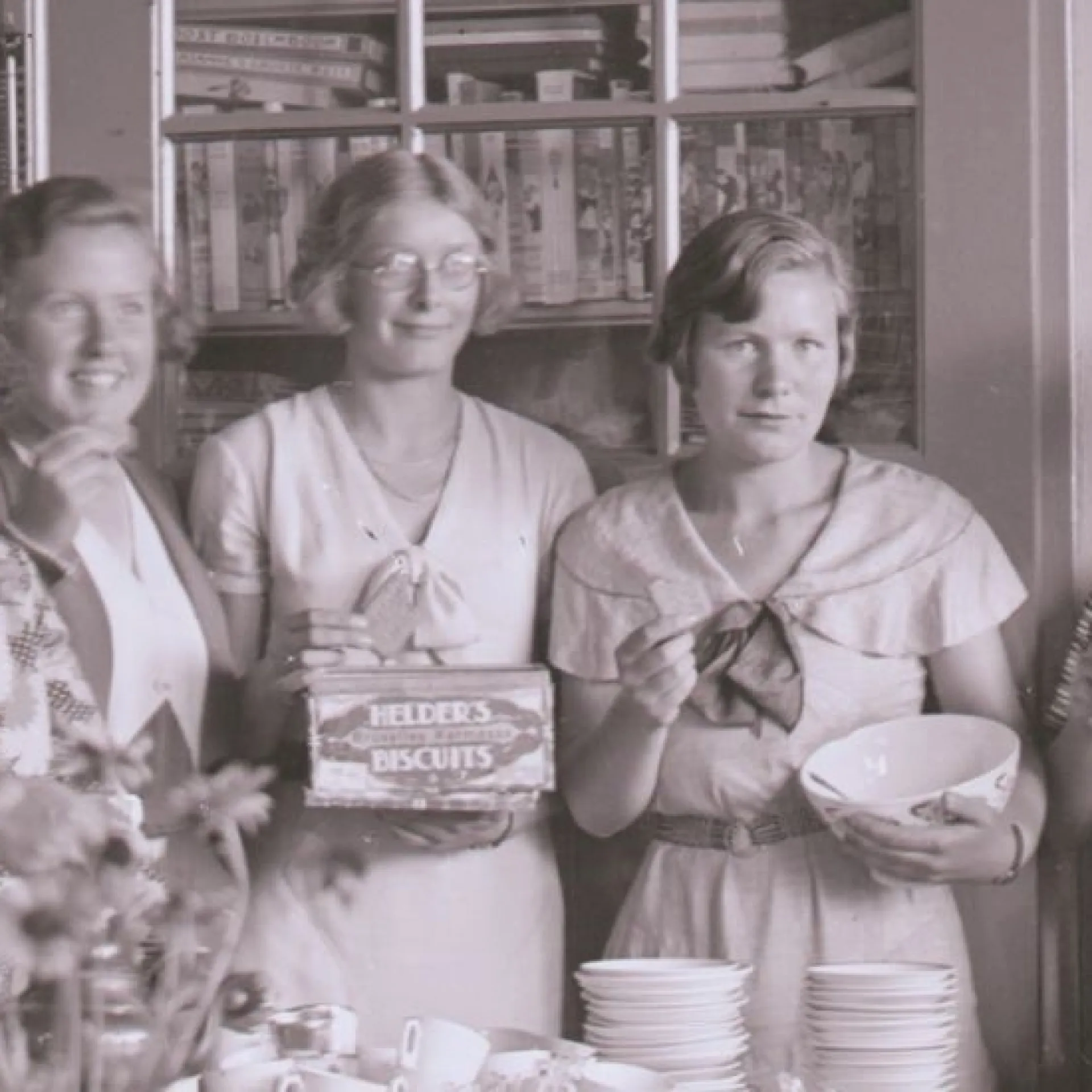 Zwart-wit foto. Groep van 8 vrouwen in klaslokaal, drinkt thee en eet biscuits. Aan de muur hangen schoolplaten. Op tafel staan stapels kopjes en schotels. 