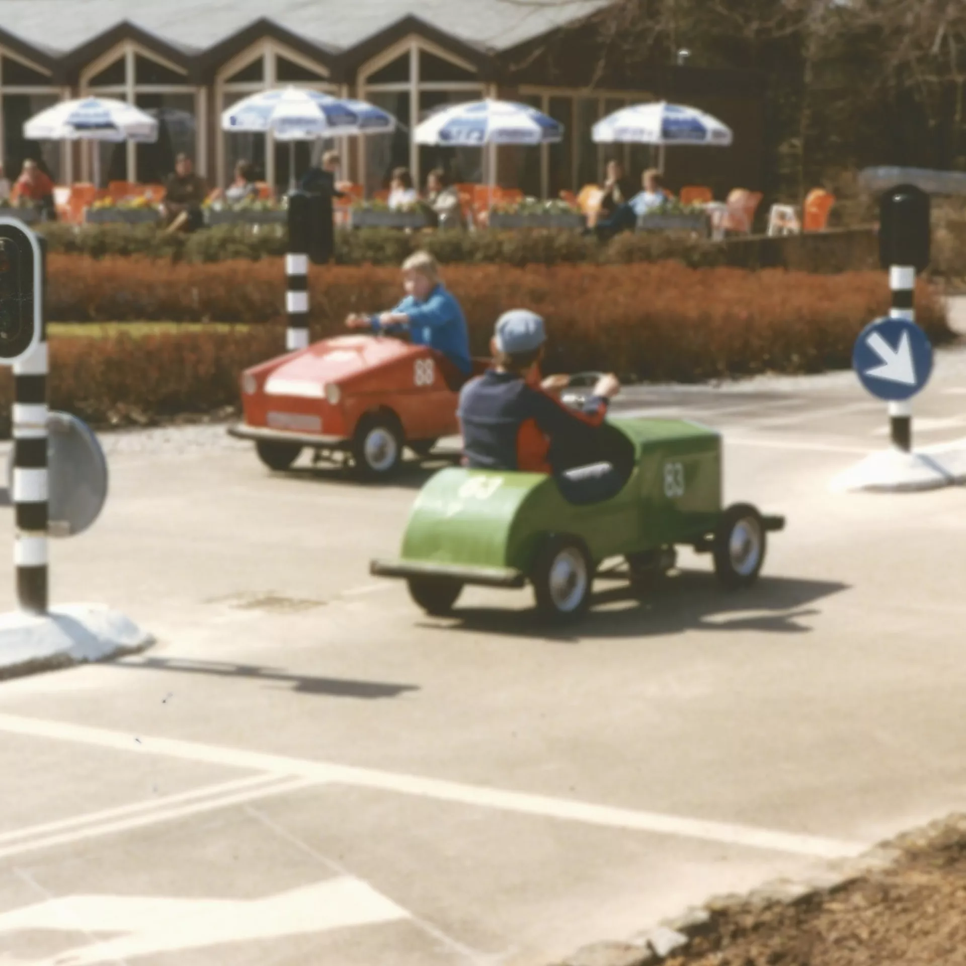 Kleurenfoto van verkeerspark Assen. Er rijden drie kleine auto's, er staan verkeerslichten. Op de achtergrond zien we een terras met parasols.