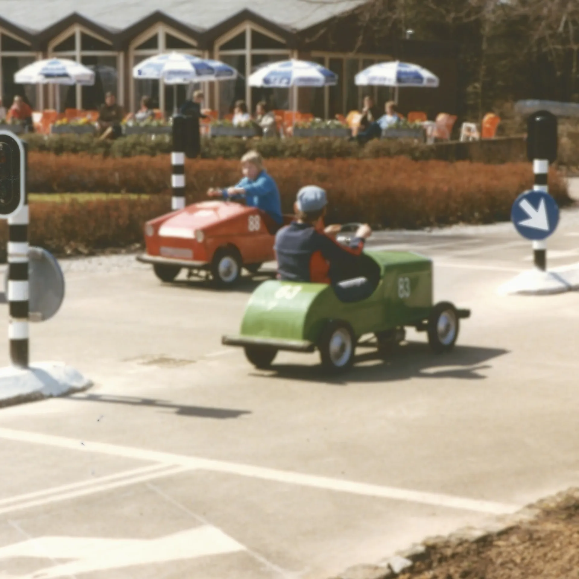 Kleurenfoto van verkeerspark Assen. Er rijden drie kleine auto's, er staan verkeerslichten. Op de achtergrond zien we een terras met parasols.